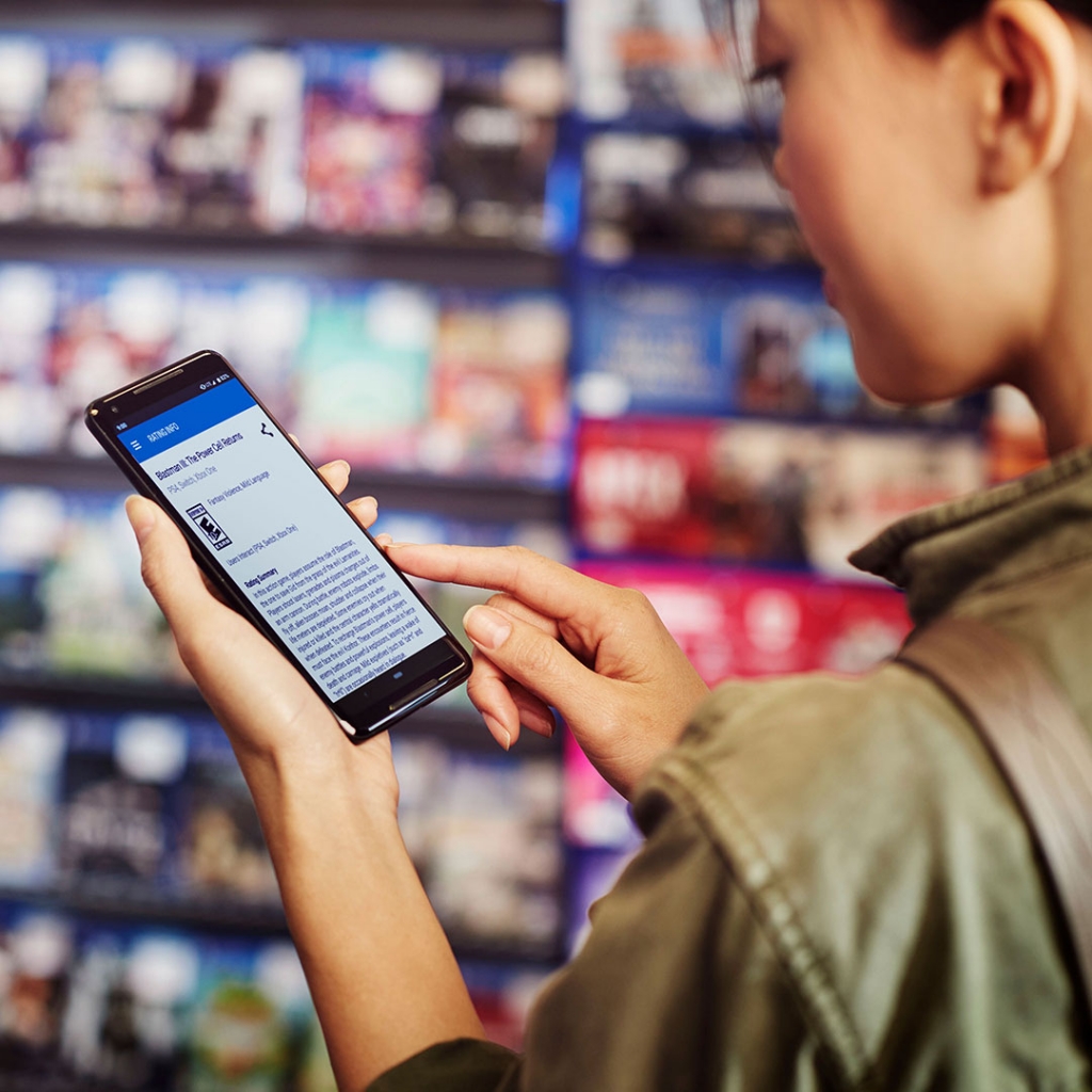 A woman in the image is browsing ESRB ratings on her smartphone, checking the age-appropriate content for video games or other media in a retail setting.